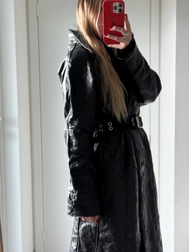May include: Black leather trench coat with a belted waist and a high collar. The coat has a textured appearance and features a double-breasted design. The coat is a long length and appears to be vintage.