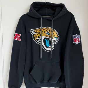 May include: Black hooded sweatshirt featuring the Jacksonville Jaguars logo on the front. The logo is a jaguar head with teal, gold, and white accents. The sweatshirt has a kangaroo pocket and the NFL and AFC logos on the sleeves.
