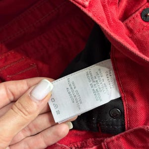 以下が含まれることがあります： 黒い内側の赤いデニムジーンズのクローズアップ。 「100％コットン」や「メキシコ製」などのテキストが表示された白いタグが見えます。 ジーンズはボタン留めと、クラシックな5ポケットデザインです。