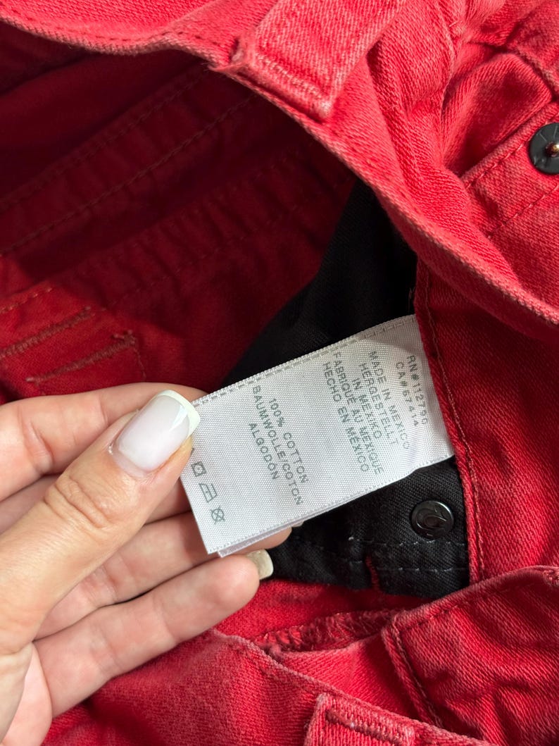 以下が含まれることがあります： 黒い内側の赤いデニムジーンズのクローズアップ。 「100％コットン」や「メキシコ製」などのテキストが表示された白いタグが見えます。 ジーンズはボタン留めと、クラシックな5ポケットデザインです。