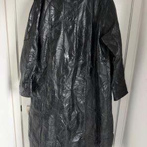 May include: A long, black leather coat with a patchwork design. The coat has a collar and long sleeves. The coat is hanging on a white door. The leather appears to be shiny.