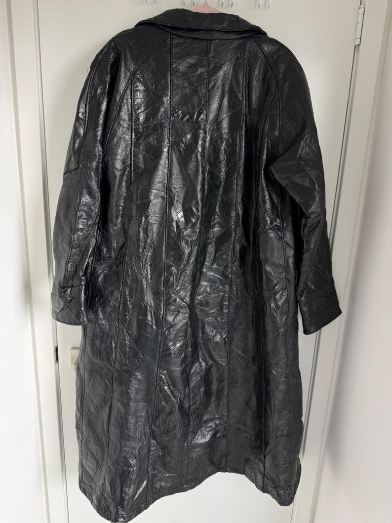 May include: A long, black leather coat with a patchwork design. The coat has a collar and long sleeves. The coat is hanging on a white door. The leather appears to be shiny.