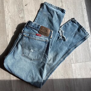 May include: Light blue denim jeans with a distressed look. The jeans feature a classic five-pocket design, a brown leather patch with the "Wrangler" logo, and a red tag. The jeans are folded on a wooden surface.