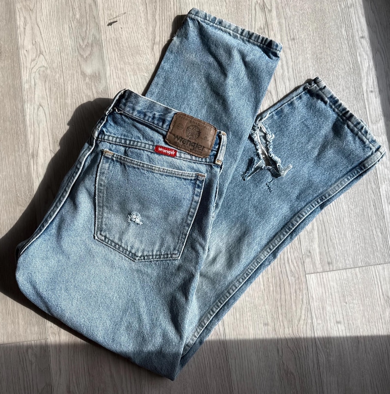 May include: Light blue denim jeans with a distressed look. The jeans feature a classic five-pocket design, a brown leather patch with the "Wrangler" logo, and a red tag. The jeans are folded on a wooden surface.