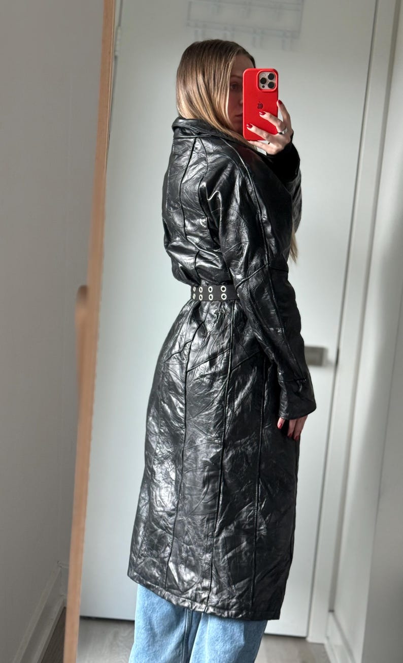 May include: A long, black, quilted leather coat with a belted waist. The coat has a high collar and a textured surface. The belt has a series of silver-colored rings. The coat is knee-length.
