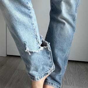 May include: Light blue denim jeans with distressed details, featuring a ripped knee on the left leg. The jeans have a classic five-pocket design and a faded wash, showing signs of wear.