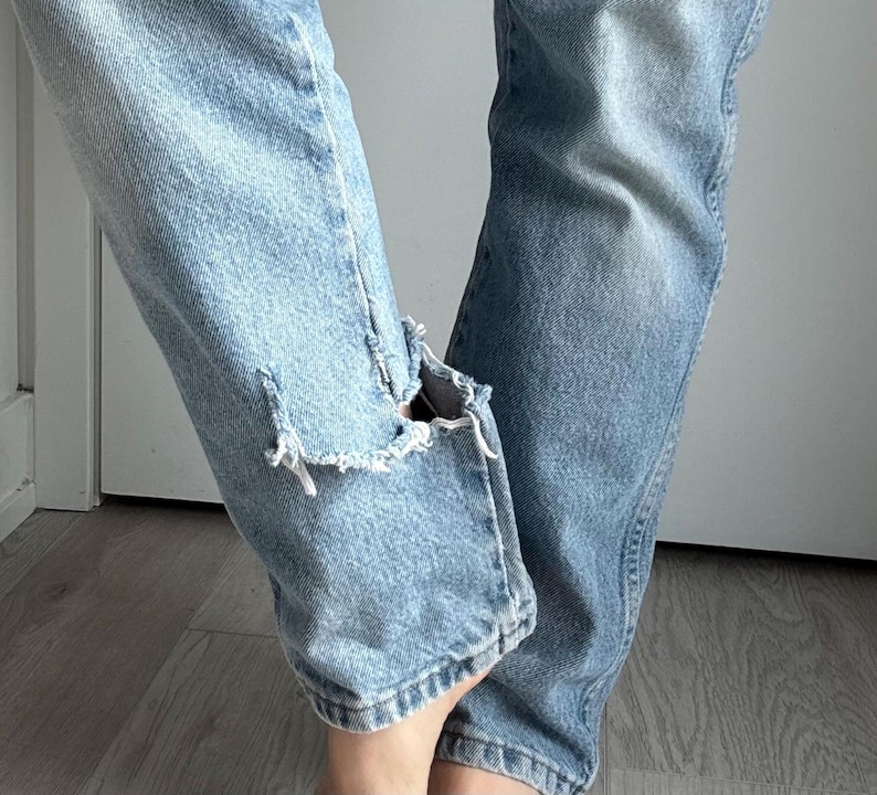 May include: Light blue denim jeans with distressed details, featuring a ripped knee on the left leg. The jeans have a classic five-pocket design and a faded wash, showing signs of wear.