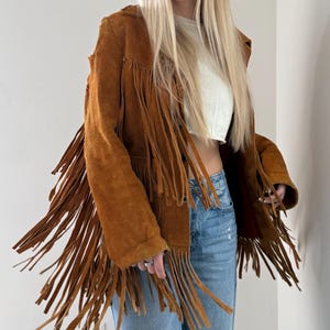 May include: A brown suede jacket with long fringe detailing. The jacket has a classic design with a button closure and a relaxed fit. The fringe adds a bohemian flair to the outerwear.