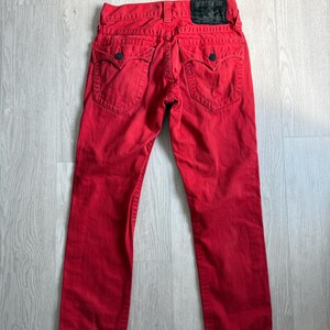 以下が含まれることがあります： クラシックな5ポケットデザインの赤いデニムジーンズ。ジーンズは、バックウエストバンドに黒い革のパッチと、バックポケットに装飾的なステッチが施されています。ジーンズは鮮やかな赤色です。