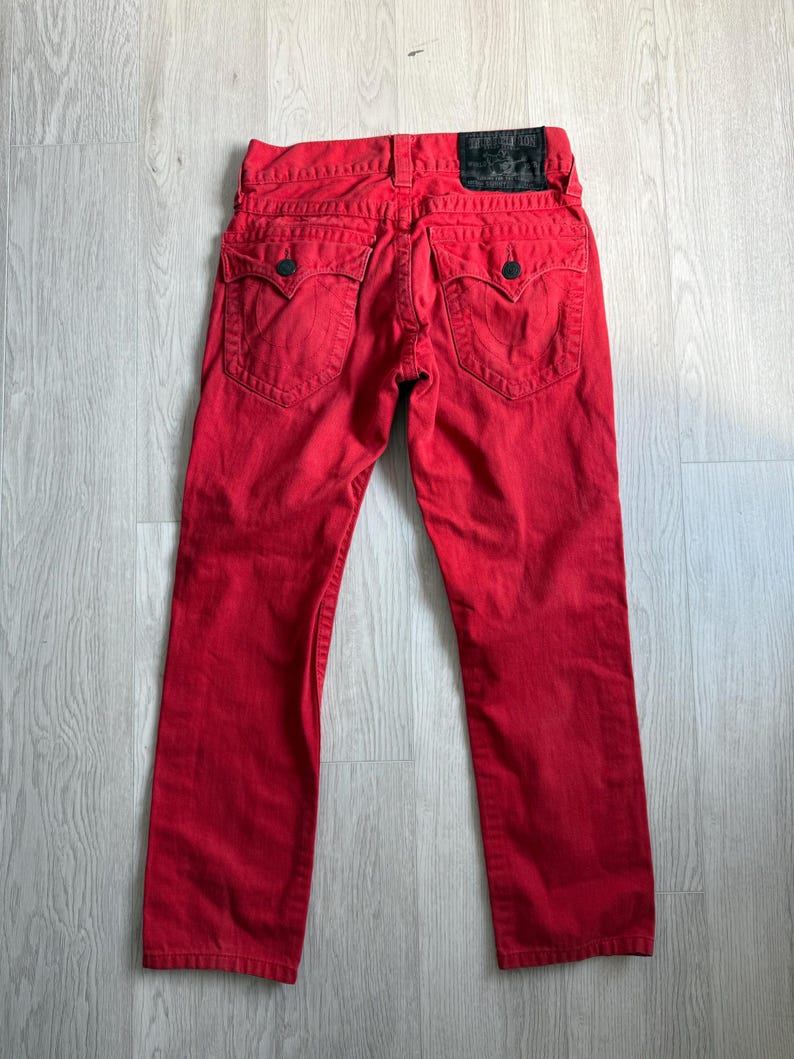 以下が含まれることがあります： クラシックな5ポケットデザインの赤いデニムジーンズ。ジーンズは、バックウエストバンドに黒い革のパッチと、バックポケットに装飾的なステッチが施されています。ジーンズは鮮やかな赤色です。