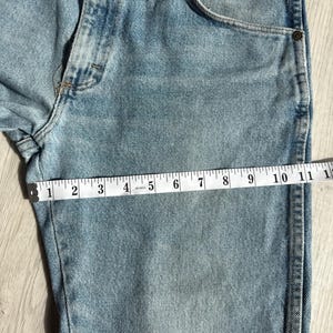 May include: Light blue denim jeans with a measuring tape showing a length of approximately 11 inches. The jeans have a classic five-pocket design and a faded wash, with visible stitching and a button closure.