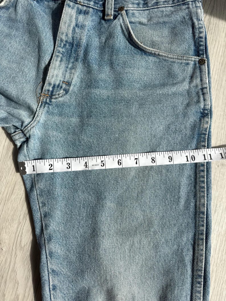 May include: Light blue denim jeans with a measuring tape showing a length of approximately 11 inches. The jeans have a classic five-pocket design and a faded wash, with visible stitching and a button closure.