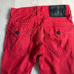 以下が含まれることがあります： True Religionブランドのラベルが見える鮮やかな赤色のデニムジーンズ。ジーンズには、装飾的なステッチと濃い青色のボタン留めが付いた2つのバックポケットがあります。ジーンズはスキニーフィットです。