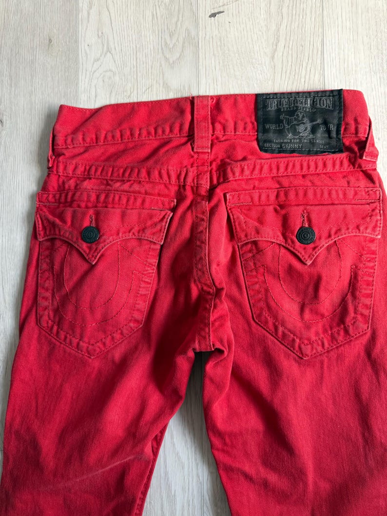 以下が含まれることがあります： True Religionブランドのラベルが見える鮮やかな赤色のデニムジーンズ。ジーンズには、装飾的なステッチと濃い青色のボタン留めが付いた2つのバックポケットがあります。ジーンズはスキニーフィットです。