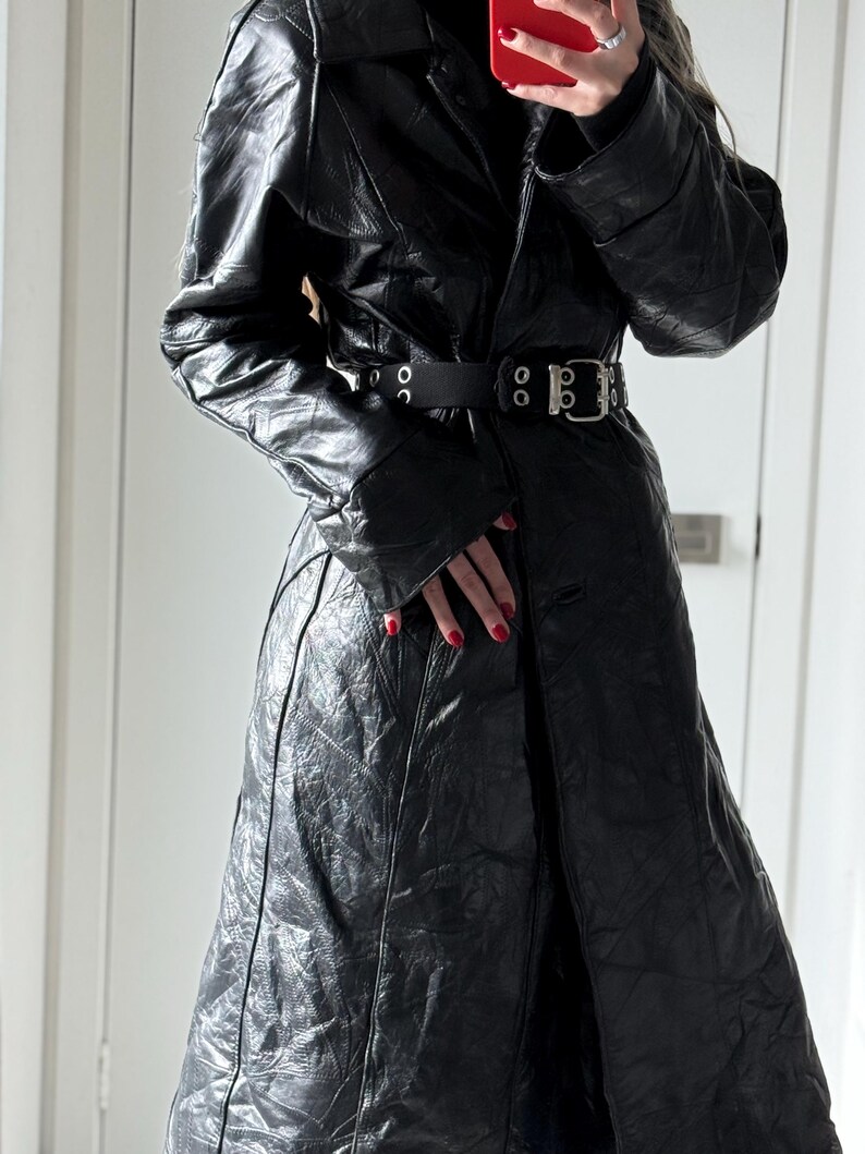 May include: A long, black leather coat with a belted waist and a notched collar. The coat has a textured surface and a split front. The belt is black with a silver buckle. The coat is a stylish outerwear garment.