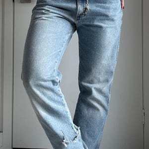 May include: Light blue denim jeans with a silver chain belt. The jeans have a distressed detail at the ankle. The person is wearing the jeans with bare feet and red nail polish.