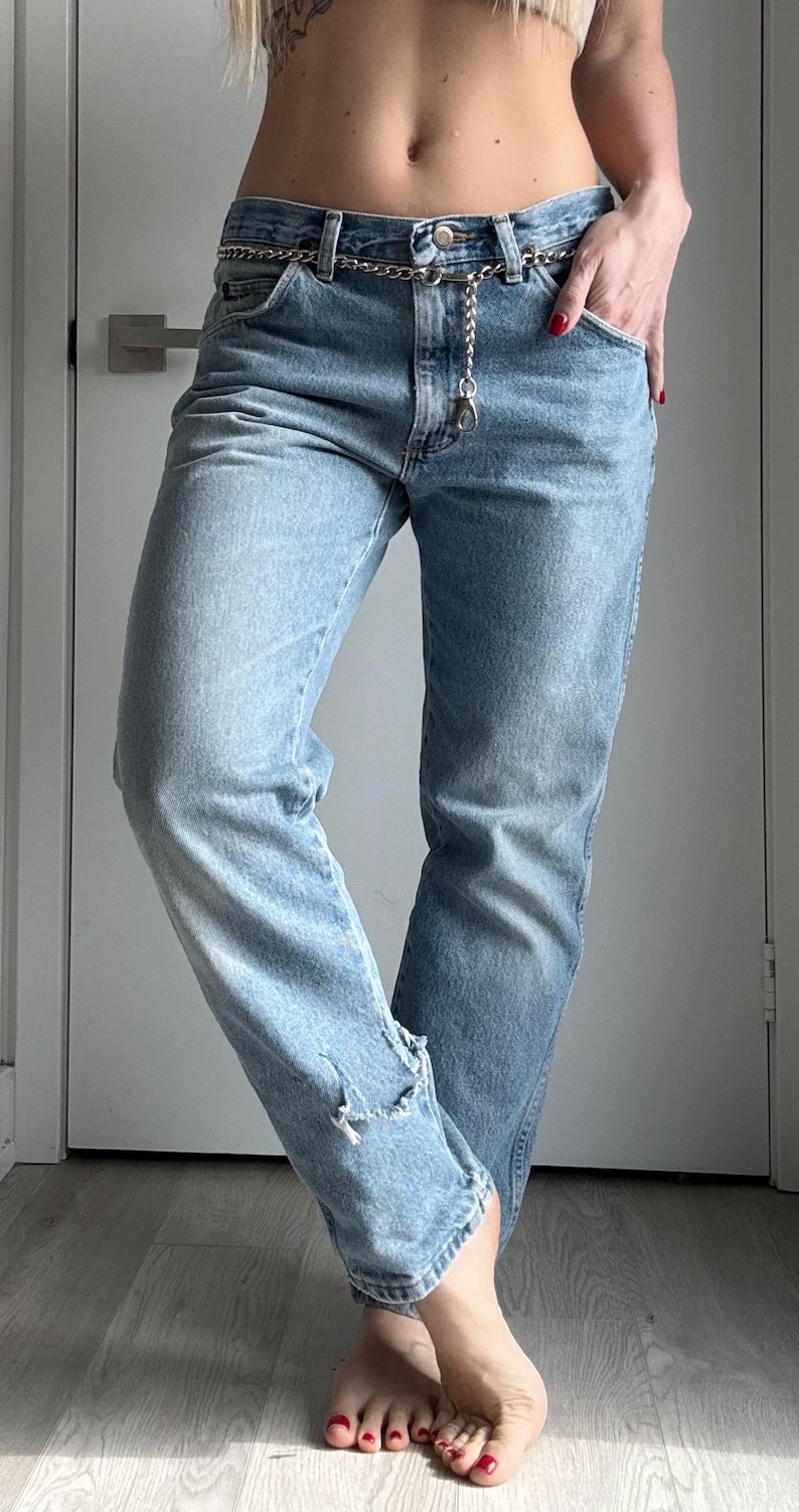 May include: Light blue denim jeans with a silver chain belt. The jeans have a distressed detail at the ankle. The person is wearing the jeans with bare feet and red nail polish.