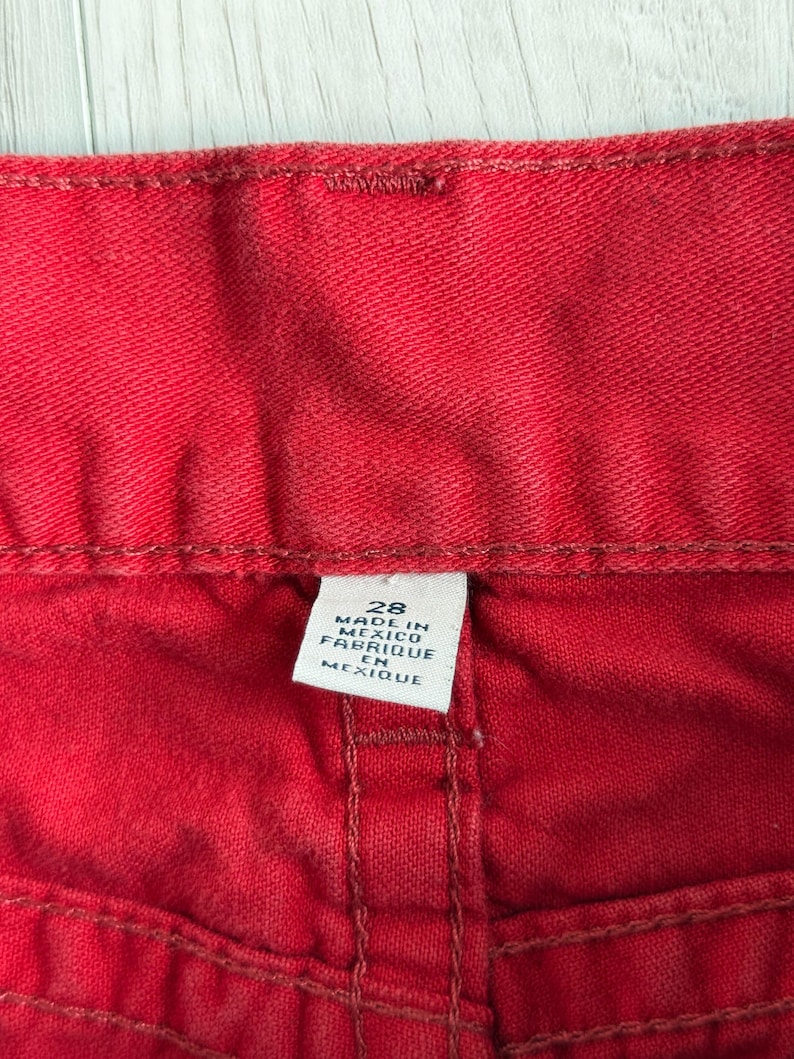 以下が含まれることがあります： 赤いデニム生地のクローズアップ。ステッチと白いタグが見えます。タグには「28 MADE IN MEXICO FABRIQUE EN MEXIQUE」と書かれています。生地はジーンズのようです。
