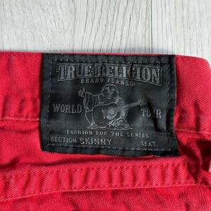 以下が含まれることがあります： 赤いデニムジーンズのクローズアップ。黒いTrue Religionブランドのラベルが付いています。ラベルにはブランド名、ギターを弾く人物のグラフィック、そして「World Tour」、「Section Skinny」、「Seat」の文字が記載されています。
