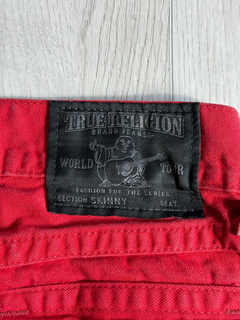 以下が含まれることがあります： 赤いデニムジーンズのクローズアップ。黒いTrue Religionブランドのラベルが付いています。ラベルにはブランド名、ギターを弾く人物のグラフィック、そして「World Tour」、「Section Skinny」、「Seat」の文字が記載されています。