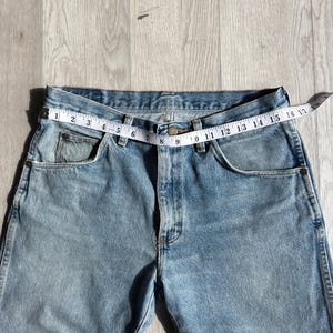 May include: Light blue denim jeans with a measuring tape across the waist. The tape reads 16 inches across the waist. The jeans have classic five-pocket styling and a button and zipper closure.