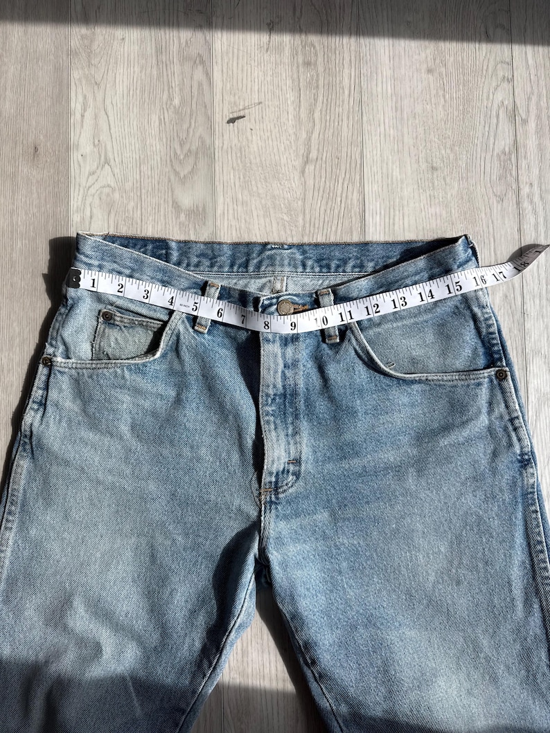 May include: Light blue denim jeans with a measuring tape across the waist. The tape reads 16 inches across the waist. The jeans have classic five-pocket styling and a button and zipper closure.