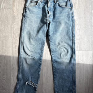 May include: Light blue denim jeans with a distressed look. The jeans have a classic five-pocket design, a button and zipper closure, and a worn area on the left leg. The jeans are laid flat on a light-colored wooden surface.