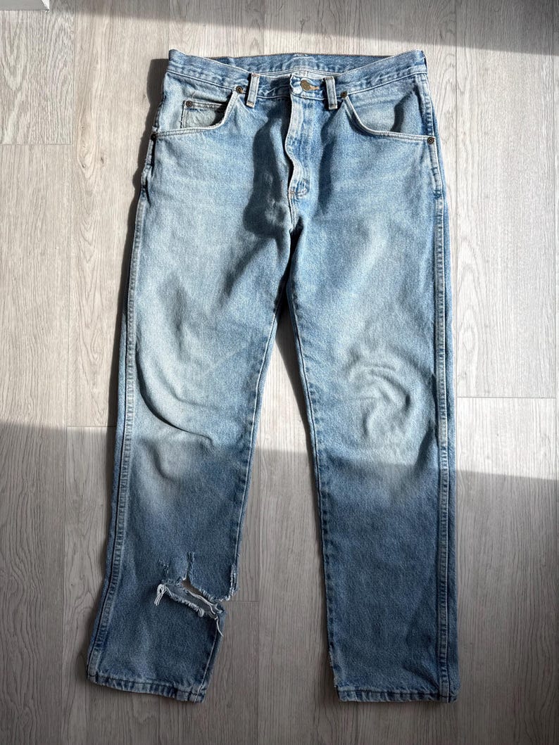 May include: Light blue denim jeans with a distressed look. The jeans have a classic five-pocket design, a button and zipper closure, and a worn area on the left leg. The jeans are laid flat on a light-colored wooden surface.