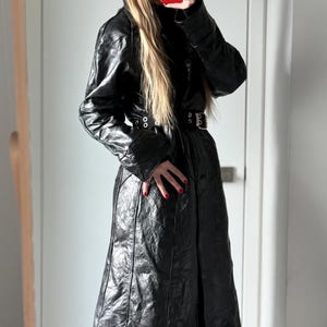 May include: A long, black leather coat with a belted waist and quilted texture. The coat features a high collar and long sleeves. The model is wearing blue jeans. The coat is a stylish outerwear piece.