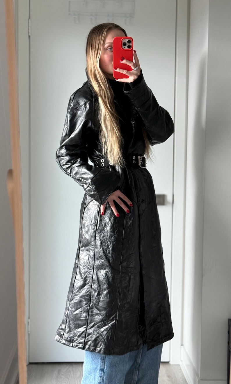 May include: A long, black leather coat with a belted waist and quilted texture. The coat features a high collar and long sleeves. The model is wearing blue jeans. The coat is a stylish outerwear piece.