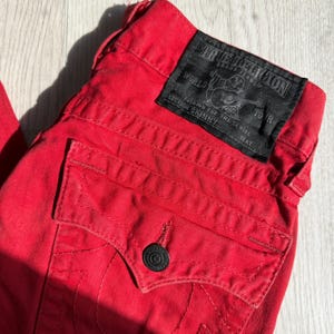 以下が含まれることがあります： 黒い革のパッチが付いた赤いデニムジーンズのクローズアップ。パッチにはブランド名とロゴが表示されています。ジーンズはボタン留めとバックポケットが付いています。ジーンズには「Section Skinny」と表示されています。