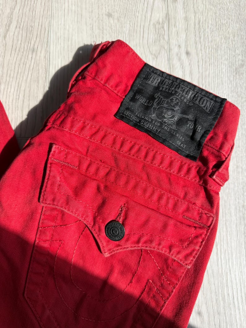 以下が含まれることがあります： 黒い革のパッチが付いた赤いデニムジーンズのクローズアップ。パッチにはブランド名とロゴが表示されています。ジーンズはボタン留めとバックポケットが付いています。ジーンズには「Section Skinny」と表示されています。