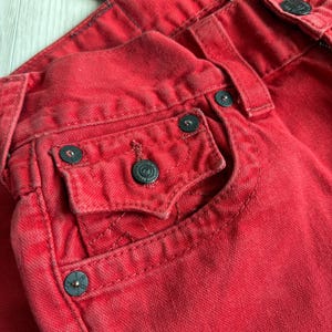 以下が含まれることがあります： 赤いデニムジーンズのクローズアップ。ジーンズは、ダークメタルボタンとリベットを備えたクラシックな5ポケットデザインが特徴です。生地はわずかに色あせた外観で、使い古された外観を示唆しています。ステッチが見え、ジーンズ全体の美しさに貢献しています。