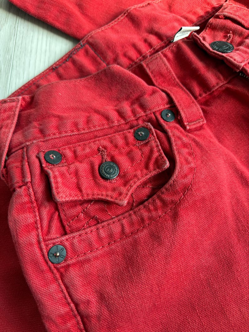 以下が含まれることがあります： 赤いデニムジーンズのクローズアップ。ジーンズは、ダークメタルボタンとリベットを備えたクラシックな5ポケットデザインが特徴です。生地はわずかに色あせた外観で、使い古された外観を示唆しています。ステッチが見え、ジーンズ全体の美しさに貢献しています。