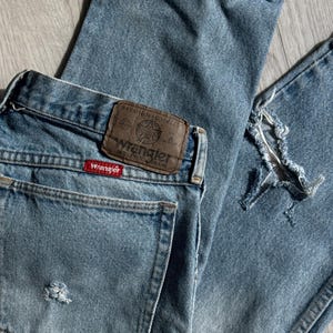 May include: Light blue Wrangler jeans with distressed details. The jeans feature a classic five-pocket design, a brown leather patch with the Wrangler logo, and a red Wrangler tag. The denim has a faded appearance.
