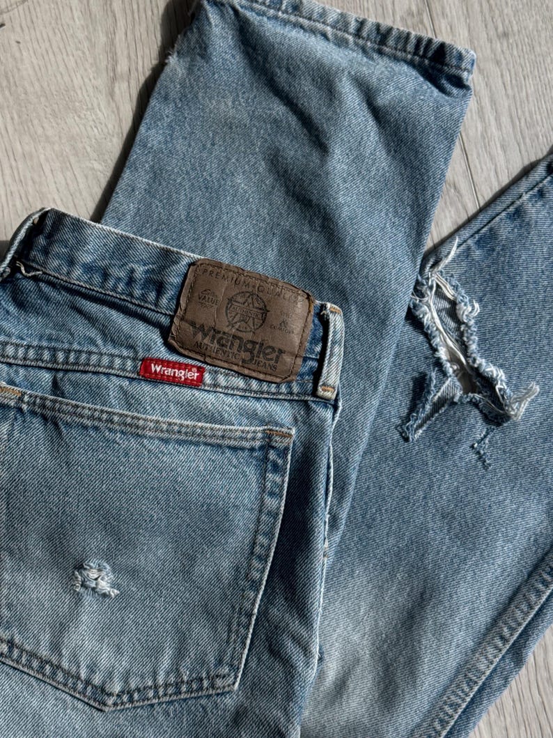 May include: Light blue Wrangler jeans with distressed details. The jeans feature a classic five-pocket design, a brown leather patch with the Wrangler logo, and a red Wrangler tag. The denim has a faded appearance.