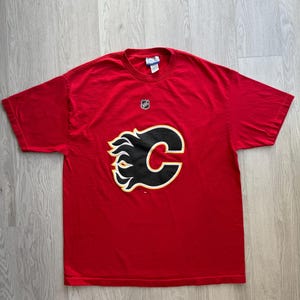 May include: A red t-shirt featuring the Calgary Flames logo. The logo is a stylized black 'C' with a flame design in black, yellow, and white. The NHL logo is visible near the neckline.
