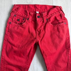 以下が含まれることがあります： 鮮やかな赤色のデニムジーンズ。クラシックな5ポケットのデザイン。ジーンズは、リベットやボタン留めなど、黒色の金具が特徴です。生地は中程度の厚さで、普段使いに適しています。ジーンズはきれいに折りたたまれ、フロントポケットとウエストバンドが強調されています。