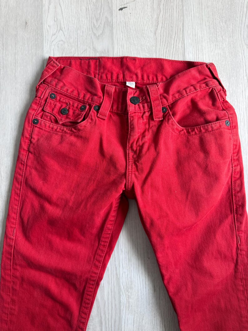 以下が含まれることがあります： 鮮やかな赤色のデニムジーンズ。クラシックな5ポケットのデザイン。ジーンズは、リベットやボタン留めなど、黒色の金具が特徴です。生地は中程度の厚さで、普段使いに適しています。ジーンズはきれいに折りたたまれ、フロントポケットとウエストバンドが強調されています。