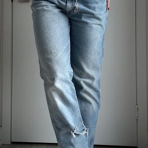 May include: Light blue denim jeans with a silver chain belt. The jeans have a distressed detail at the knee. The model is wearing the jeans with their hand in the pocket. The jeans are a classic, casual style.
