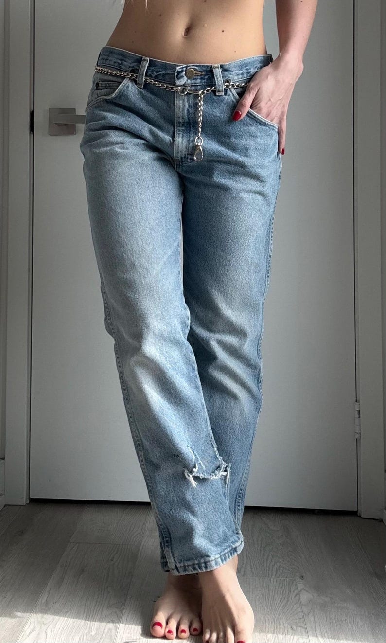 May include: Light blue denim jeans with a silver chain belt. The jeans have a distressed detail at the knee. The model is wearing the jeans with their hand in the pocket. The jeans are a classic, casual style.