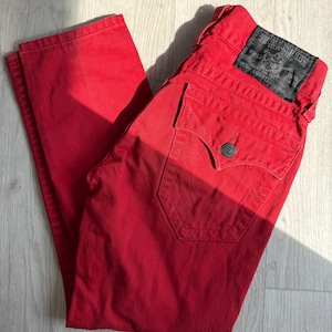 May include: Bright red denim jeans with a classic five-pocket design. The jeans feature a black leather patch with white text and a button closure. The jeans are folded and lying on a light-colored wooden surface.