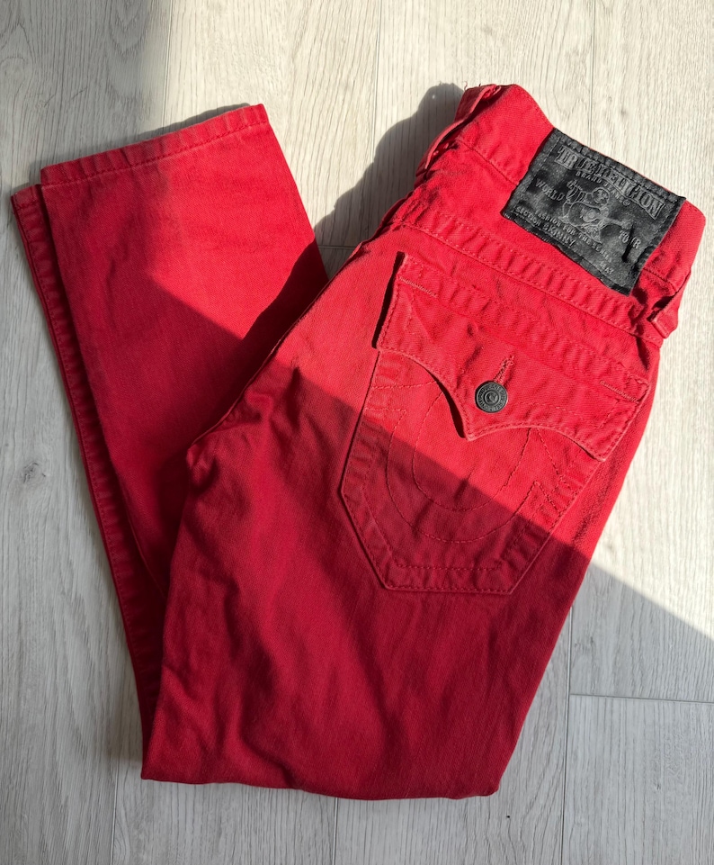 以下が含まれることがあります： 鮮やかな赤色のデニムジーンズ。クラシックな5ポケットデザイン。ジーンズは、白い文字が入った黒い革のパッチとボタン留めが特徴です。ジーンズは明るい色の木製の表面に折りたたまれています。