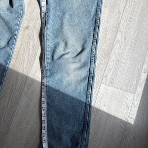 May include: Light blue denim jeans with a measuring tape running along the leg. The tape shows measurements in inches. The jeans are laid out on a light-colored wooden surface.