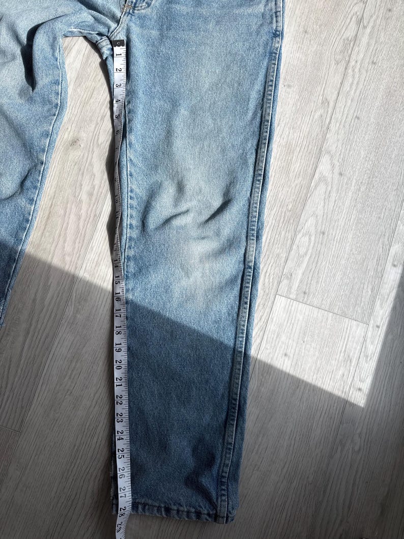 May include: Light blue denim jeans with a measuring tape running along the leg. The tape shows measurements in inches. The jeans are laid out on a light-colored wooden surface.