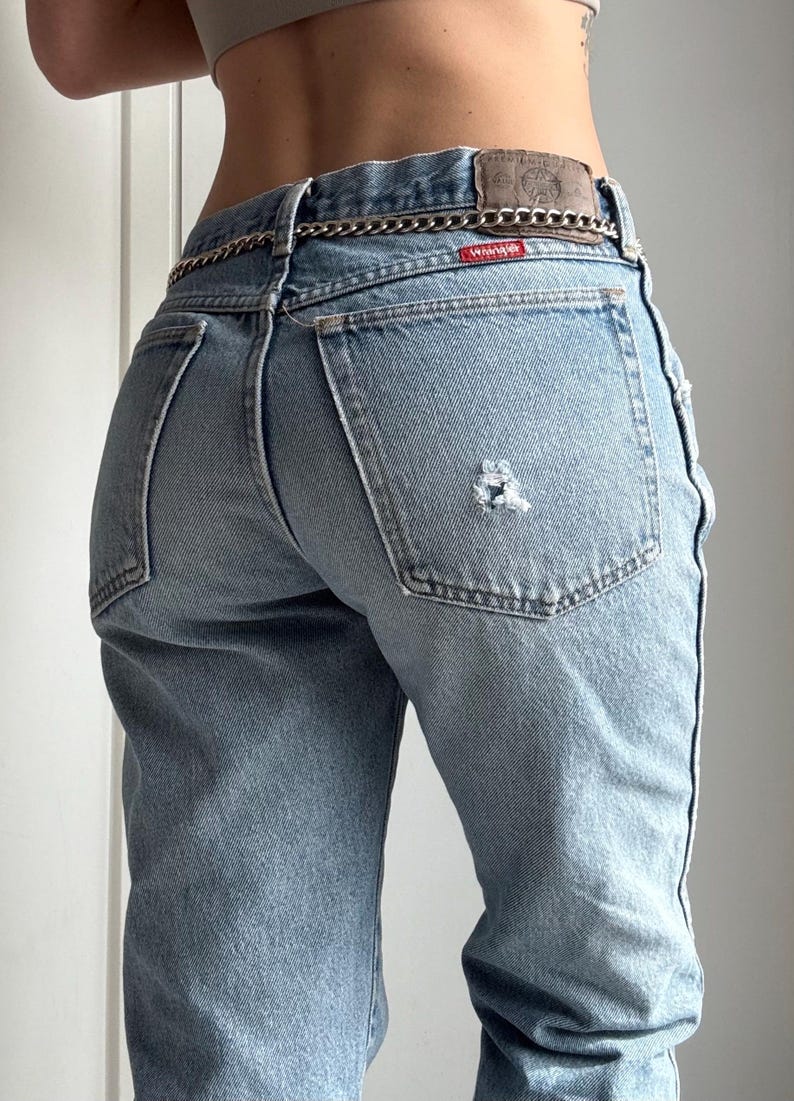 May include: Light blue denim jeans with a distressed patch on the back pocket. The jeans have a high waist and a silver chain belt. The Wrangler brand label is visible on the back.