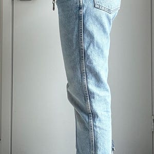 May include: Light blue denim jeans with a silver chain belt. The jeans have a classic five-pocket design and a straight leg fit. The denim is faded, and the jeans have a distressed detail at the ankle.