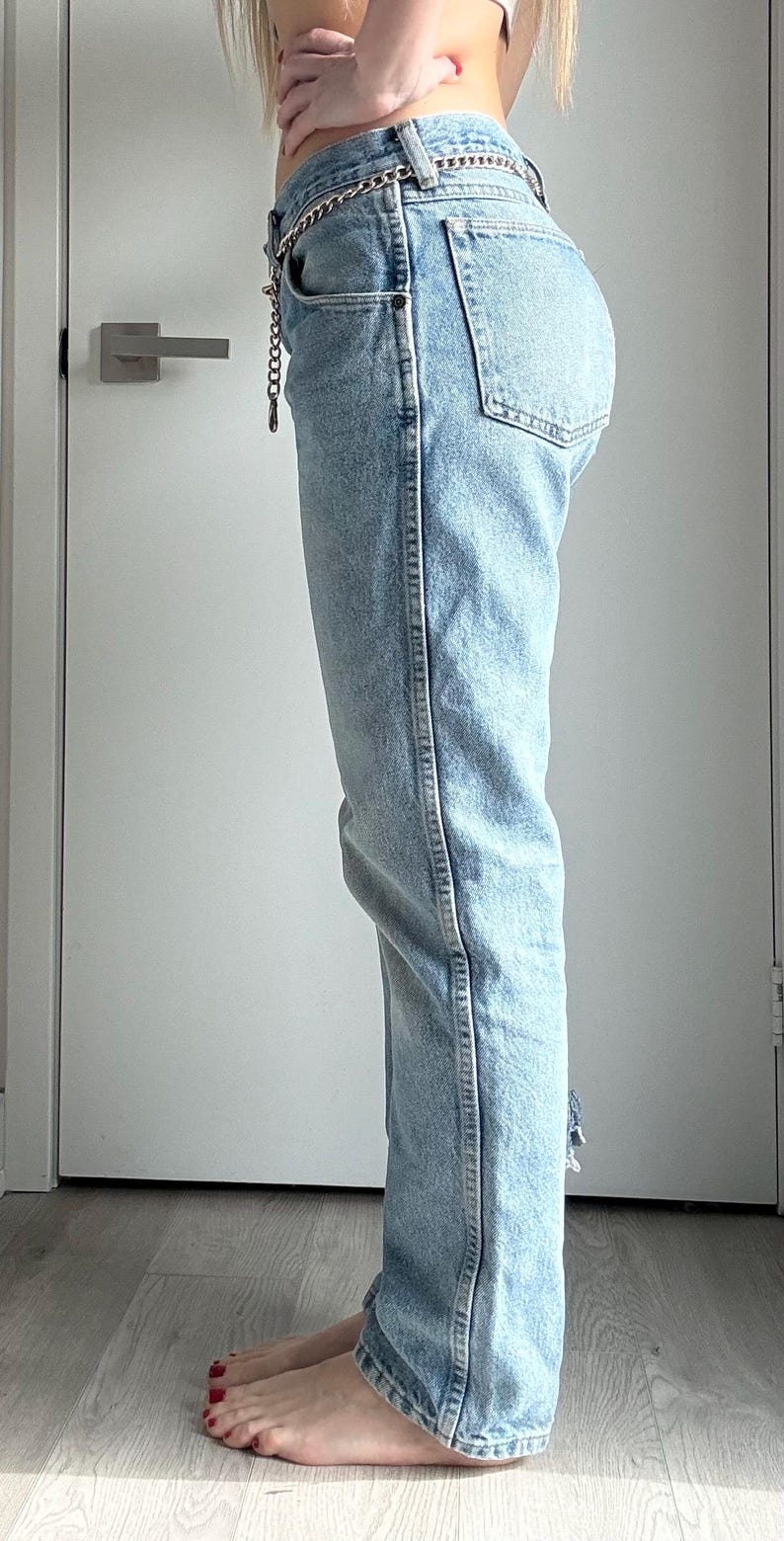 May include: Light blue denim jeans with a silver chain belt. The jeans have a classic five-pocket design and a straight leg fit. The denim is faded, and the jeans have a distressed detail at the ankle.