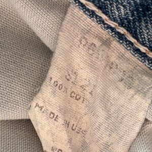 May include: Close-up of a denim jeans label. The label is off-white with black text that reads "SIZE 32", "100% COT", and "MADE IN USA". The jeans are blue with white stitching.