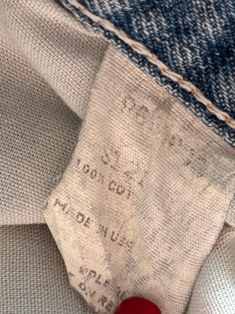 May include: Close-up of a denim jeans label. The label is off-white with black text that reads "SIZE 32", "100% COT", and "MADE IN USA". The jeans are blue with white stitching.